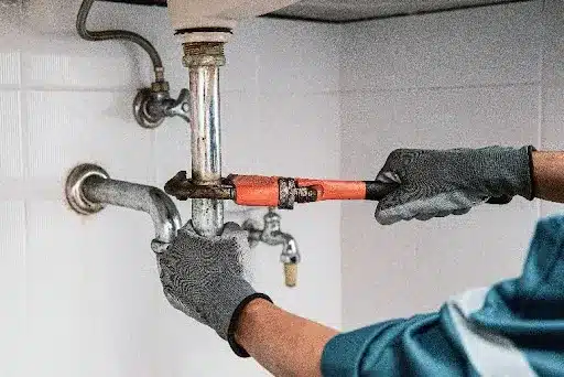 A plumber wearing grey gloves and holding a wrench to dislodge a clog from a drain in Riverside, MO. A drain cleaning service in a residential bathroom.