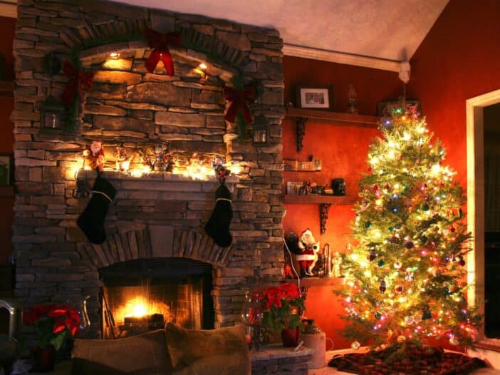 Large decorated Christmas tree next to a large brick fireplace & holiday decor in the back with a dark red wall