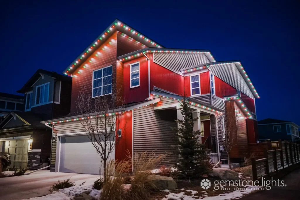 A suburban house with Gemstone Lights gracefully outlining the roof against a night sky. Snow covers parts of the yard and driveway, adding a serene touch to the winter scene.