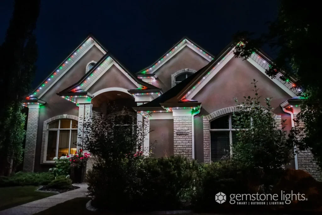 The house exterior glows at night with a roofline illuminated by red, green, and white lights, showcasing the elegant touch of Gemstone Lights. The Gemstone Lights logo is subtly placed in the bottom right corner.