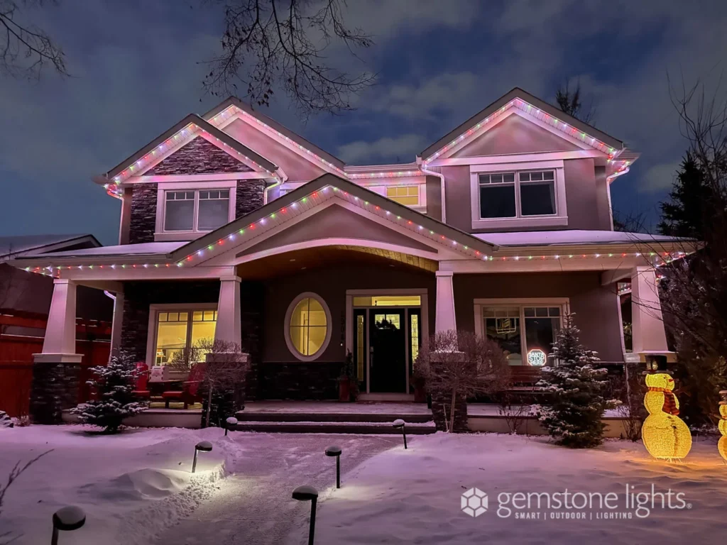 A two-story house is adorned with Gemstone Lights in a rainbow of colors. Snow blankets the ground, and a charming snowman sculpture stands proudly in front.