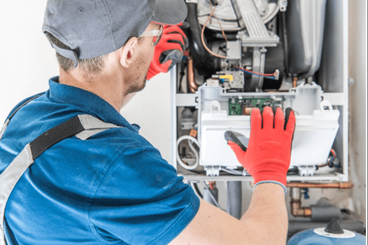 A technician wearing red gloves and a cap works on a boiler's internal components, holding a circuit board.