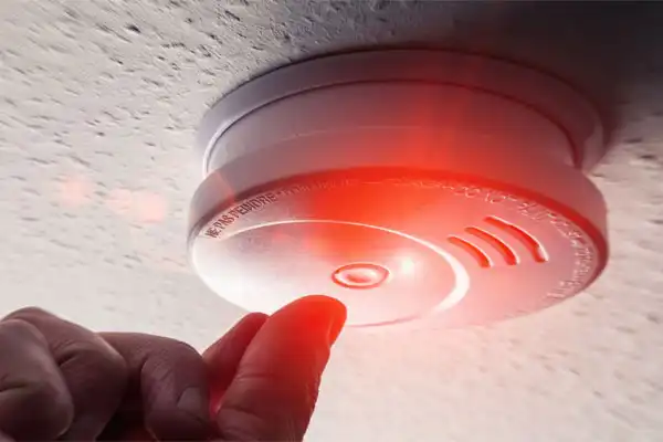 a combination smoke & carbon monoxide (CO) detector beeping a red alert light while a homeowner reach's to reset the detector