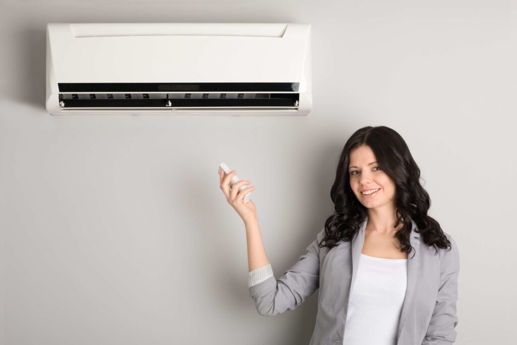 Woman standing near a wall-mounted air conditioner, holding a remote control, wearing a gray blazer and white shirt, smiling.