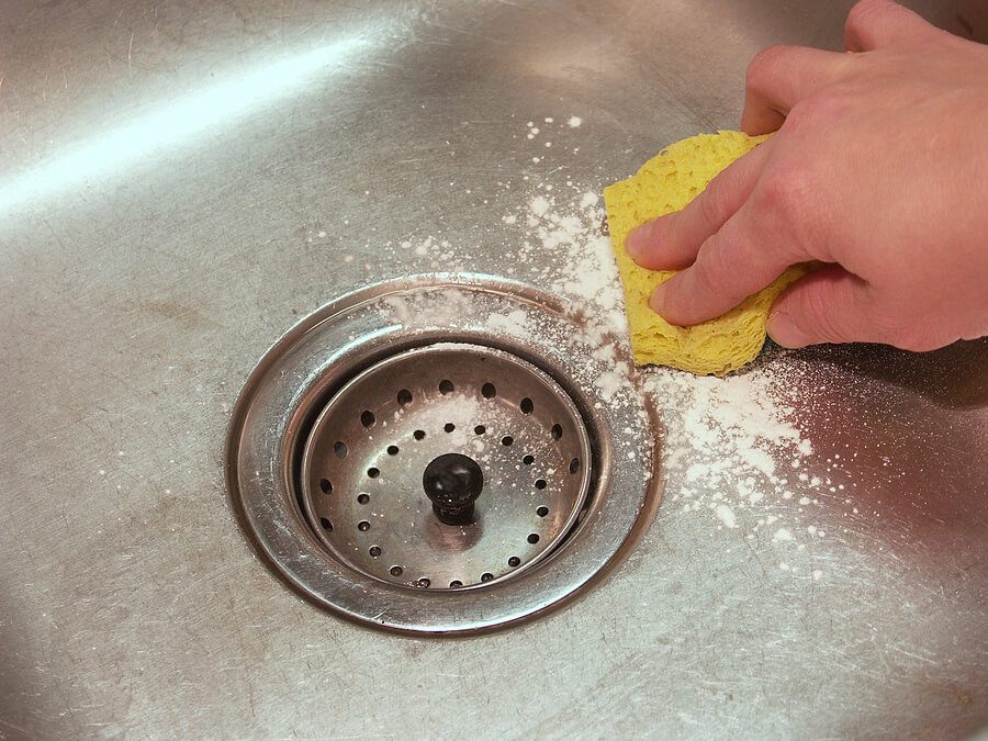 Hand scrubbing a stainless steel sink with a yellow sponge and cleaning powder near the drain.