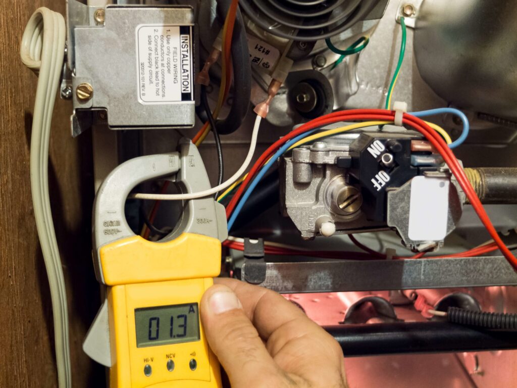 A person uses a clamp meter to measure current in a metal junction box with wires and mechanical components. The digital display reads 0.13.