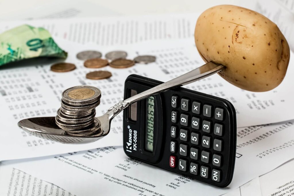 A spoon with coins and a potato balance on a calculator over scattered documents and currency.
