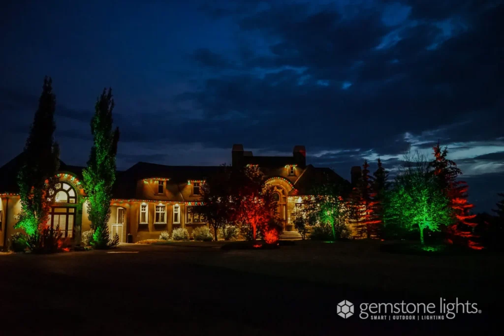 A large house at night, adorned with Gemstone Lights that cast a vibrant palette of red, green, and warm yellow hues. Trees and bushes bask in this radiant glow as the dark sky looms above with scattered clouds, creating an enchanting nighttime spectacle.