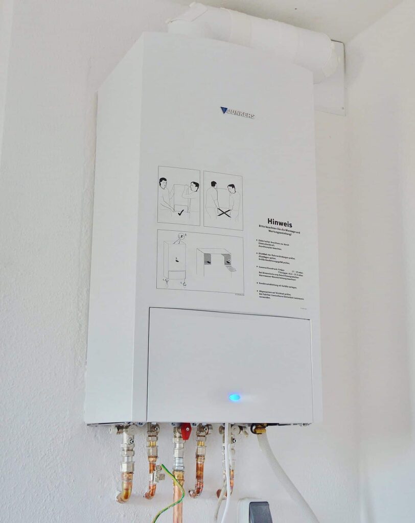 A wall-mounted white water heater with various pipes connected at the bottom. It has instructional illustrations and text on its front panel.
