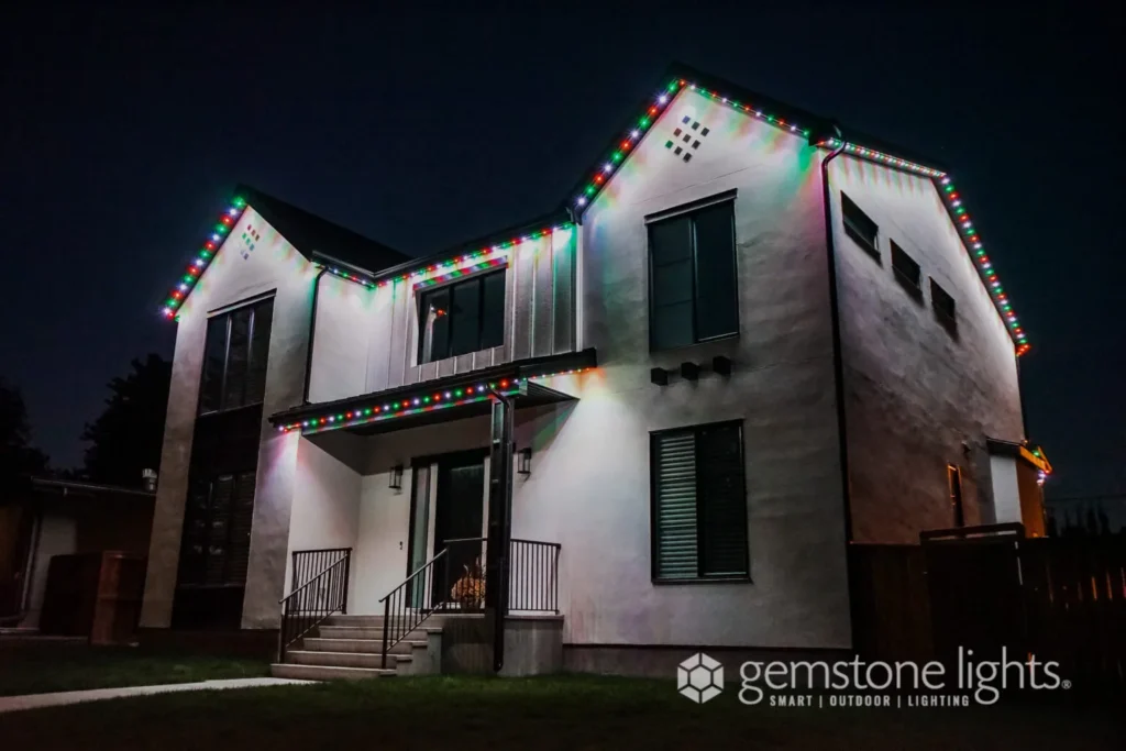 A two-story house radiantly shines with multicolored Christmas lights along the roofline, courtesy of Gemstone Lights. It is nighttime, and the "Gemstone Lights" logo proudly illuminates the scene, enhancing the festive ambiance.