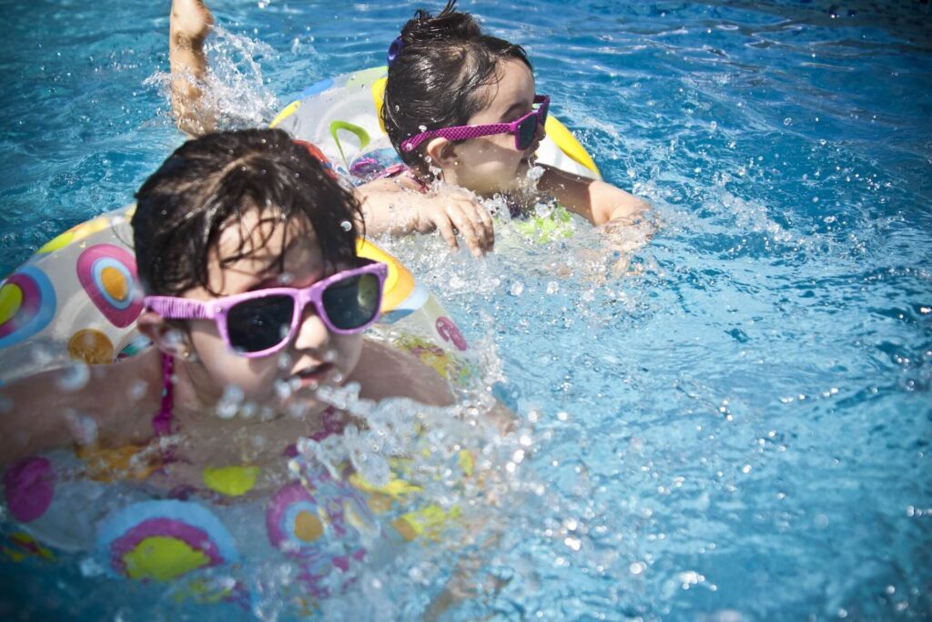 Two children in colorful inflatable rings and sunglasses play in a swimming pool, splashing water and enjoying a sunny day.