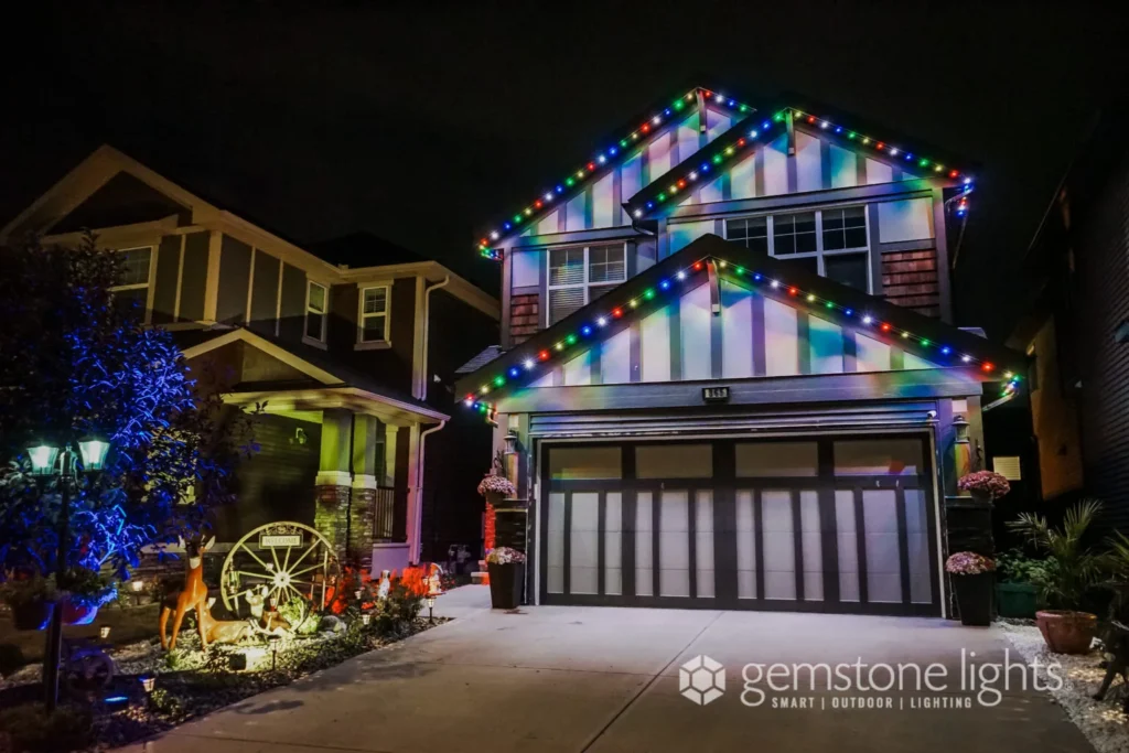 House with colorful Christmas lights on the roof and garage, a lit pathway, and a garden wheel decoration. The festive scene shines bright with Gemstone Lights, featuring their logo on the bottom right.