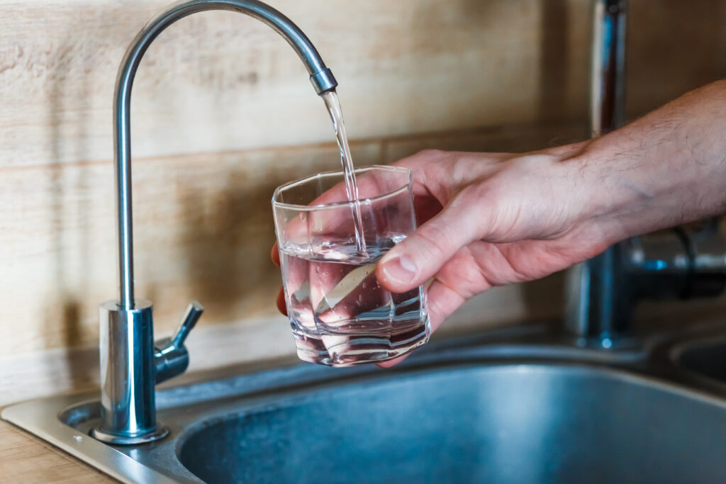 A person fills a glass with water from a kitchen faucet over a stainless steel sink.