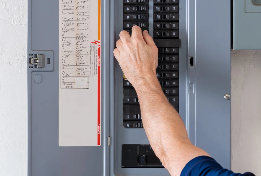 A person adjusts a switch in a circuit breaker panel with multiple labeled switches in a metal enclosure.